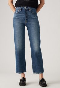 Kõrge vöökoht, sinised denim püksid sirgete jalgadega, kerge kulumine ja õmblustega detailid taskutel, koos mustade loafersitega.