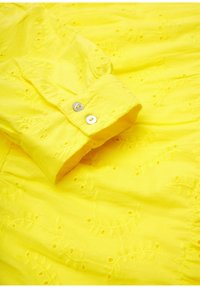 Żółta bawełniana bluzka z haftowanymi wzorami liści i małymi detalami z oczkami. Zapięcie na guzik z mankietami i dwoma srebrnymi guzikami. Gładka tekstura.