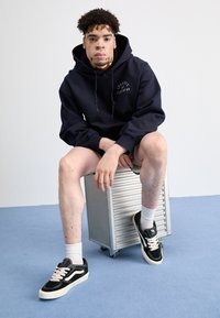 Navy hoodie with "Class of 89" logo, black sneakers with white accents, worn with shorts, sitting on a metallic stool on a blue floor.