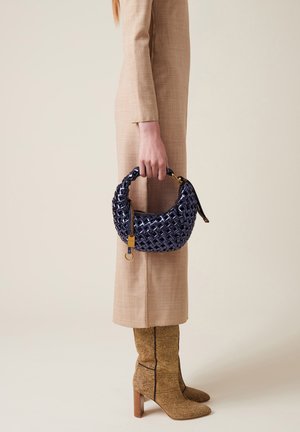 Borsa intrecciata blu navy con dettagli dorati tenuta in una mano. La borsa ha una forma arrotondata e un manico strutturato. Abito beige texturizzato e stivali al ginocchio con motivo.