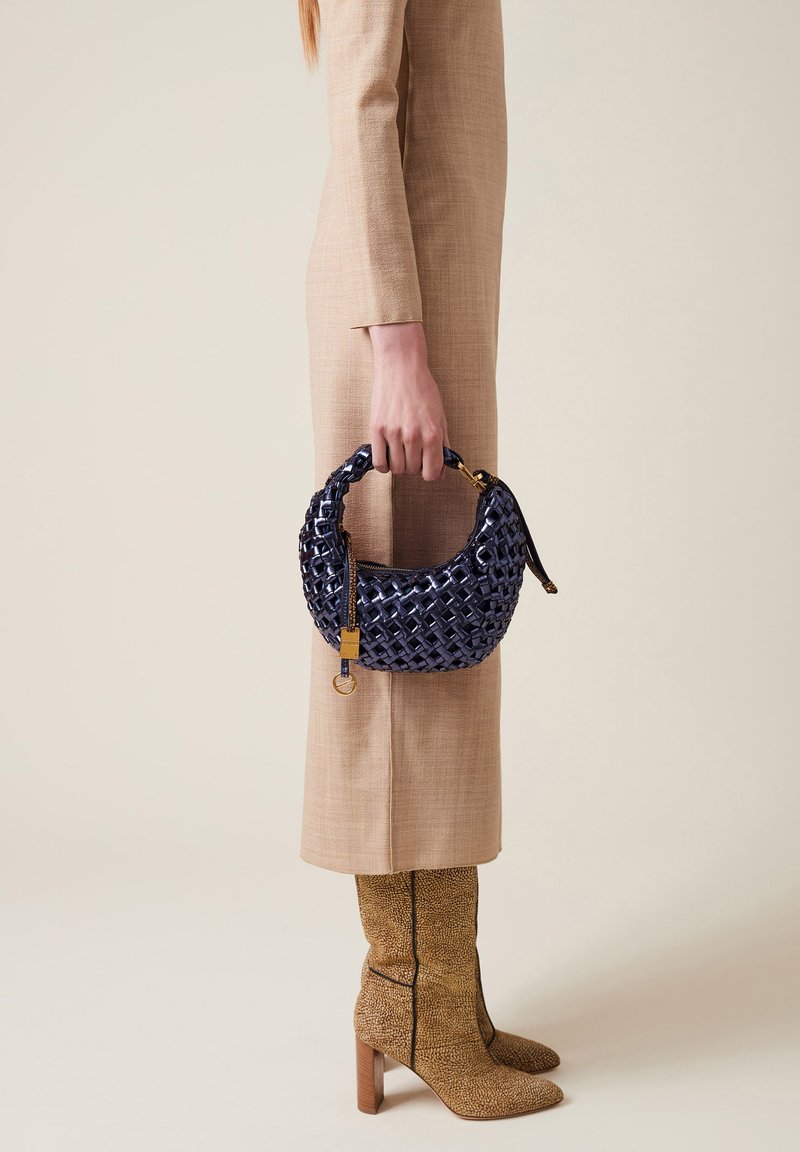Gewebte marineblaue Handtasche mit goldenen Akzenten, die in einer Hand gehalten wird. Die Tasche hat eine runde Form und einen stabilen Griff. Beigefarbenes, strukturiertes Kleid und gemusterte knielange Stiefel.