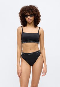 Γυναίκα με σγουρά μαλλιά που φοράει μαύρο bralette Calvin Klein και εσώρουχο ψηλόμεσο, ποζάροντας μπροστά σε μια απλή ανοιχτόχρωμη επιφάνεια, φορώντας γυαλιά ηλίου.