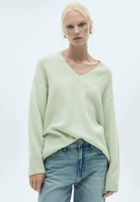 Hellgrüner V-Ausschnitt-Pullover aus weichem, strukturiertem Strickstoff. Breite Ärmel und lockere Passform, kombiniert mit blauen Jeans.