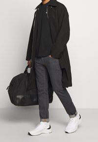 Black long coat, black t-shirt, grey jeans, white sneakers with black accents, holding a black duffel bag. Simple, modern design elements.