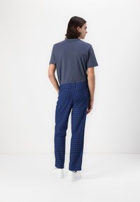 Mladić s kosom do ramena, obučen u tamnosivu majicu kratkih rukava, plave karirane hlače i bijele tenisice, stoji ispred bijelog zida.