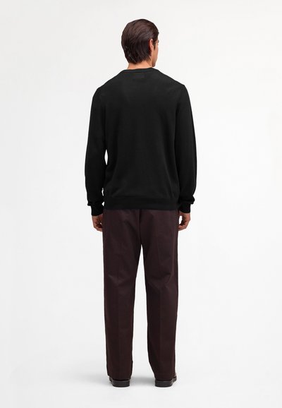 Mand stående med ryggen til iført en sort langærmet skjorte og mørkebrune bukser mod en hvid baggrund.