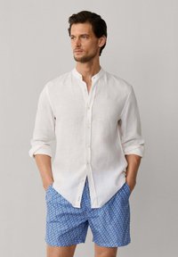 Homme portant une chemise blanche à col mandarin avec les manches retroussées et un short bleu à motifs, debout les mains dans les poches.