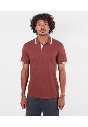 Homme aux cheveux bouclés portant un polo marron à manches courtes avec un liseré blanc et un pantalon foncé, debout devant un fond clair uni.