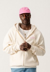 Cream zip-up hoodie with front pockets and pink "BLKYS" text. Worn over a white shirt, paired with light blue jeans and a pink cap.