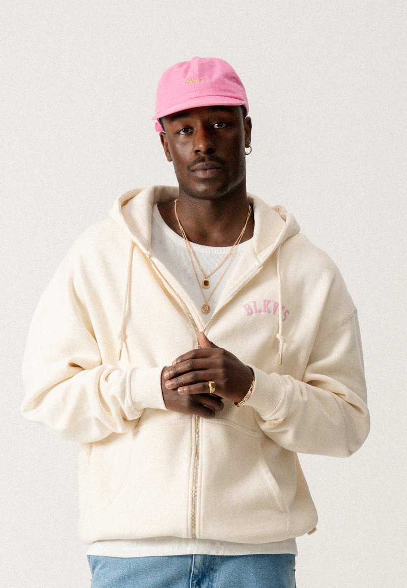 Cream zip-up hoodie with front pockets and pink "BLKYS" text. Worn over a white shirt, paired with light blue jeans and a pink cap.