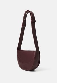 Burgundská kožená crossbody taška s zakriveným tvarom, nastaviteľným remienkom a minimalistickým dizajnom. Obsahuje hladký povrch a jemné švy.