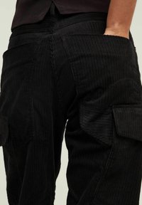 Zwarte corduroy broek met brede, uitgesproken ribbels, voorzien van zijzakken en een gestructureerde afwerking die duurzaamheid en functionaliteit benadrukt.