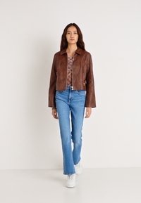 Chaqueta corta de cuero marrón con cuello, combinada con jeans acampanados azul claro y zapatillas blancas. La modelo está de pie contra un fondo neutro.