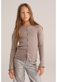 Ein junges Mädchen mit langen Haaren trägt eine knopfgebundene taupefarbene gerippte Strickjacke und glänzende silberne Hosen und steht vor einem schlichten beigefarbenen Hintergrund.