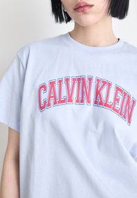 Helles blaues Baumwoll-T-Shirt mit fettem roten und blauen "CALVIN KLEIN"-Text in einem gewölbten Design. Kurze Ärmel und runder Ausschnitt.
