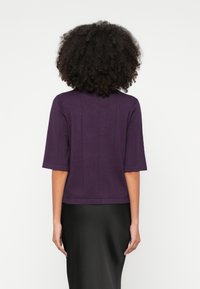 Violeta, īsroku tops ar augstu apkakli, teksturētu audumu un vertikālām šuvju detaļām; kombinēts ar gludu melnu svārku.