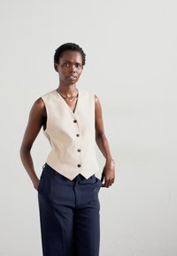 Vesta bej, fără mâneci, cu decolteu în V și cinci butoni negri, asociată cu pantaloni largi bleumarin. Material neted cu o croială ajustată.