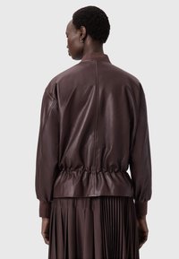 Hnědá kožená bomber bunda s elastickým pasem, spadlými ramenními švy a hladkou texturou. Zřetelná plisovaná sukně pod ní.