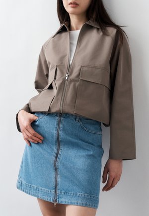 Frau trägt eine kurze taupefarbene Jacke mit großen Klappentaschen und einem Reißverschluss vorne, kombiniert mit einem hellblauen Denim-Minirock mit Reißverschluss vorne.