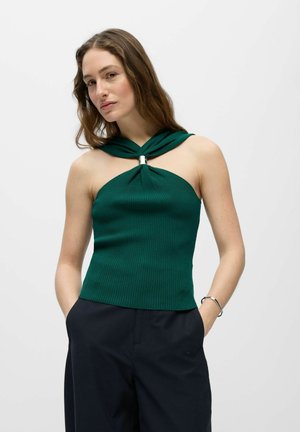 Femme portant un haut vert sans manches côtelé avec un col torsadé et un pantalon noir, debout les mains dans les poches devant un fond uni.