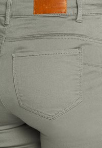 Grå bomullsblandade byxor med en bakficka, med en rektangulär läderpatch på midjan och sömnadsdetaljer längs sömmarna.