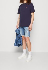 Marinblå t-shirt med en liten logotyp, kombinerad med ljusa denimshorts, vita sneakers och en vikd denimjacka som visar slitna detaljer.