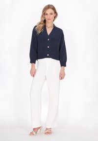 Cardigan corto blu navy con scollatura a V e cinque bottoni dorati, abbinato a pantaloni bianchi a gamba larga e sandali con tacco a blocco di colore chiaro.