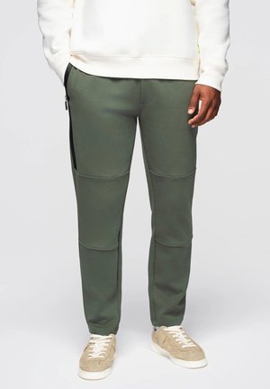 Groene sweatpants van zacht materiaal, met zijzakken met rits, gestikt detail en een taps toelopend ontwerp. Gecombineerd met lichte sneakers.