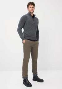 Grauer melierter Strickpullover mit einem halbseitigen Reißverschlusskragen, kombiniert mit olivgrünen Chinos und schwarzen Stiefeletten. Eingekrempelte karierte Bündchen sind sichtbar.