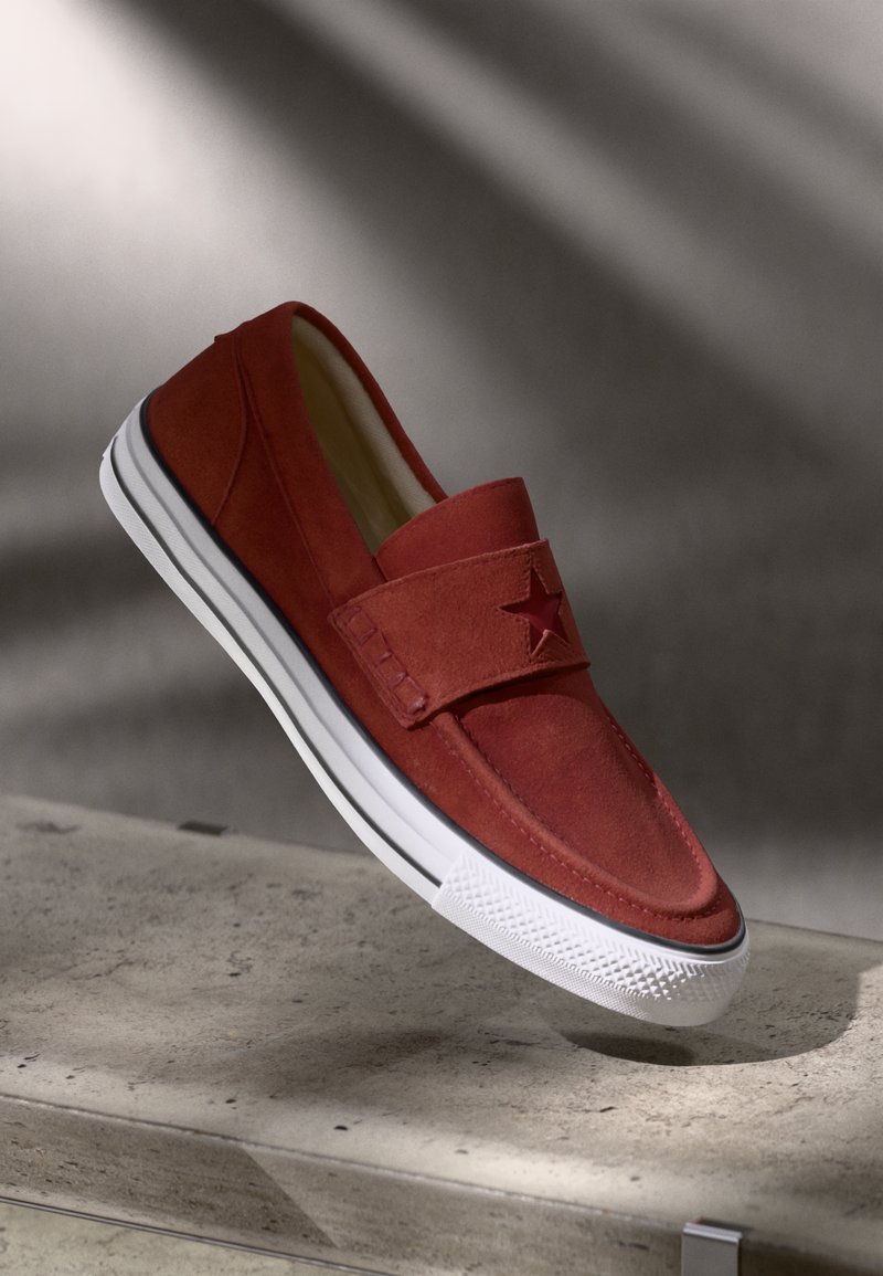 Sapato slip-on em camurça vermelha com um acento de estrela, sola de borracha branca e riscas pretas contrastantes. Textura suave, design de perfil baixo.