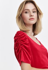 Rotes Samtoberteil mit V-Ausschnitt und Puffärmeln mit Raffung. Der Stoff hat eine weiche Textur und eine glatte Oberfläche.