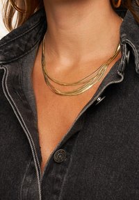 Gouden gelaagde ketting met dunne, golvende strengen tegen een donkerdenim shirt. Bevat een close-up van textuur en ontwerpelementen.