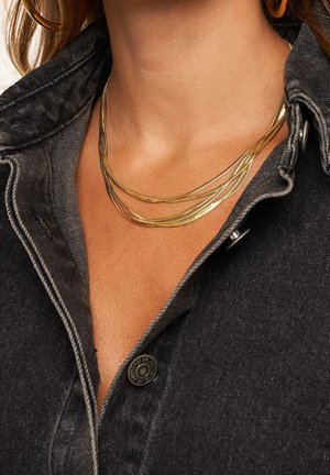 Gouden gelaagde ketting met dunne, golvende strengen tegen een donkerdenim shirt. Bevat een close-up van textuur en ontwerpelementen.