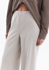 Pantaloni beige chiaro con una vestibilità comoda, dotati di una cintura elastica e tasche laterali, abbinati a una camicia marrone con bottoni.