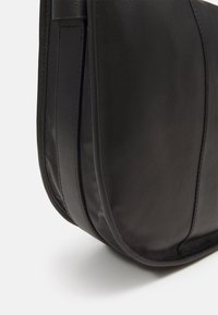 Melnas ādas plecu soma ar noapaļotu formu, gludu tekstūru un smalku dūrienu detaļām sānos.