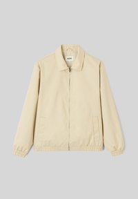 Veste beige à fermeture éclair avec col, manches longues, poignets et ourlet élastiques, et deux poches latérales sur fond gris clair uni.