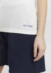 Biela bavlnená tričko s modrou výšivkou "OXMO" a detailom srdca, spárované s tmavomodrými šortkami. Hladká textura, okrúhly výstrih.
