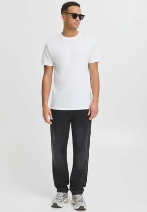 Hvit t-skjorte med korte ermer, svarte jeans og grå sneakers. Modellen har på seg mørke solbriller og står foran en nøytral bakgrunn.