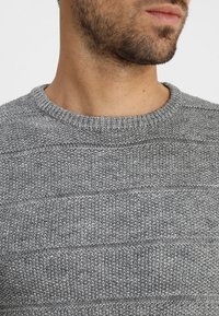 Pull en maille grise avec un motif texturé, présentant des rayures horizontales et un col côtelé. Le matériau semble doux et respirant.