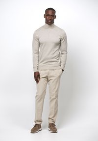 Beiger Rollkragenpullover, hellfarbige, gefaltete Hose und braune Sneakers. Porträt eines stehenden Mannes mit neutralem Hintergrund.