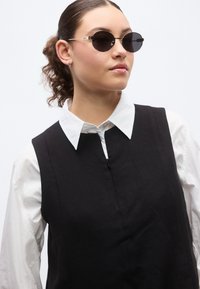 Στρογγυλά μαύρα γυαλιά ηλίου με λεπτές χρυσές μεταλλικές λεπτομέρειες. Το μοντέλο φοράει ένα μαύρο αμάνικο φόρεμα πάνω από ένα λευκό πουκάμισο με γιακά. Καθαρές γραμμές και μινιμαλιστικός σχεδιασμός.