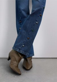 Jeans acampanados de mezclilla con detalles de cuentas coloridas en el dobladillo, combinados con zapatos de tacón de cuero marrón con punta puntiaguda. Materiales texturizados visibles.