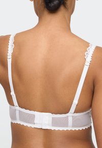 Widok z tyłu osoby noszącej biały biustonosz z koronkową lamówką, regulowanymi ramiączkami i zapięciem na trzy haftki na neutralnym tle.