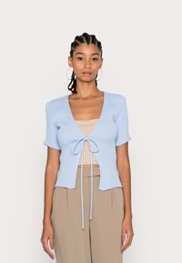 Suéter cano azul claro com acabamento em nervuras, com mangas curtas, decote em V profundo e fecho de laço na frente. Usado sobre uma blusa bege e combinado com calças beges.