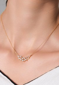 Złoty naszyjnik z kwiatowym wzorem ozdobiony przezroczystymi, szlifowanymi w kształt marquise i okrągłymi kamieniami. Łańcuszek jest delikatny i spoczywa na obojczyku.