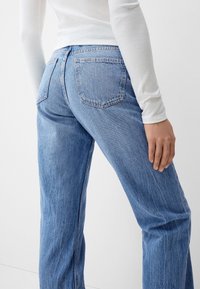 Lichtblauwe denimjeans met een rechte pijp, twee achterzakken en subtiele vervaging, gecombineerd met een wit longsleeve shirt.