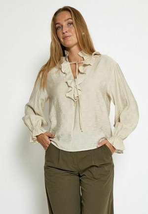 Beige blouse with ruffled neckline, textured fabric, and long sleeves with ruffled cuffs, paired with olive green trousers.