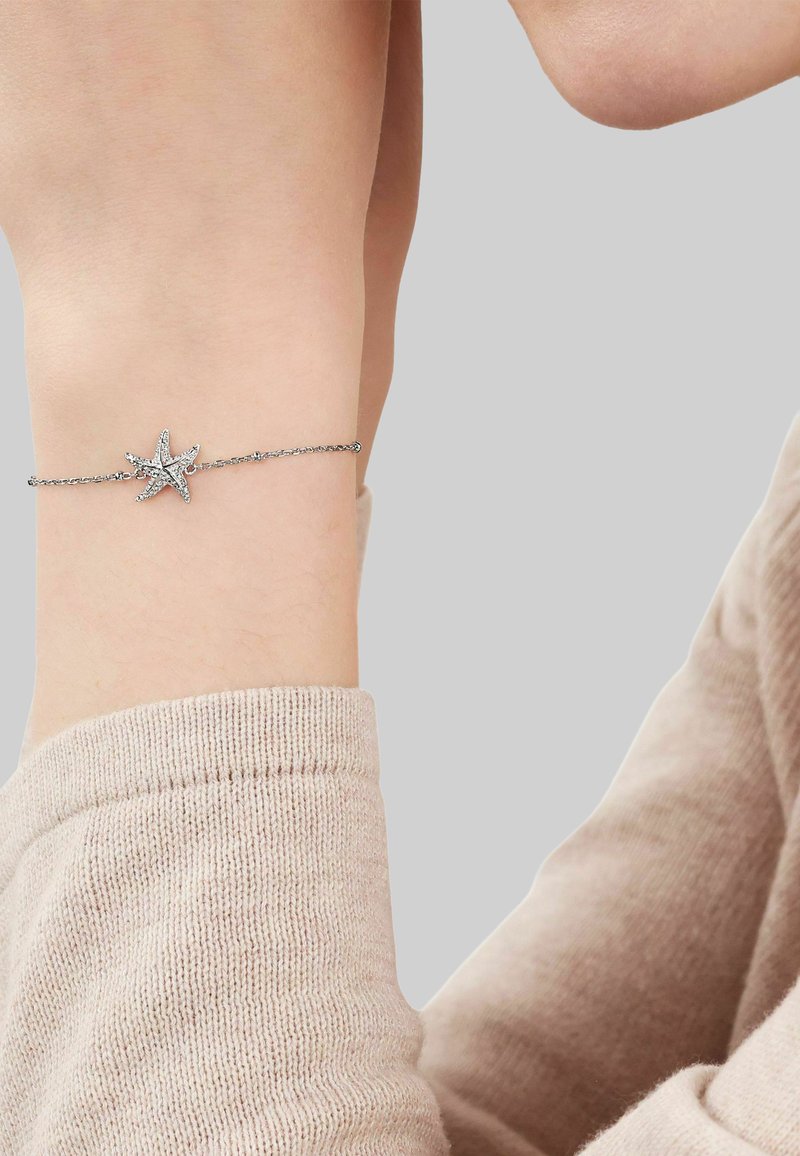 Pulsera con encanto de estrella de mar plateada en una muñeca, llevada sobre una prenda de manga larga de punto color beige claro contra un fondo gris liso.
