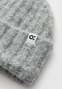 Grijze gebreide beanie met een structuurpatroon. Heeft een klein zwart-wit label met "OX" aan de zijkant. Zacht, harig materiaal.