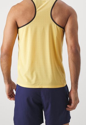 Hombre con camiseta deportiva sin mangas amarilla con pequeñas perforaciones y pantalones cortos azul marino, visto desde atrás sobre un fondo liso.